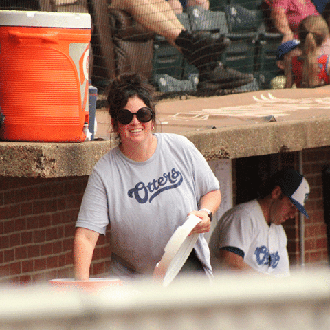 A white woman wearing large round sunglasses and a light gray “Otters” T-shirt is smiling while holding a stack of cups in a baseball dugout. A man in a matching shirt and hat is seated behind her, and an orange cooler is visible on the ledge.