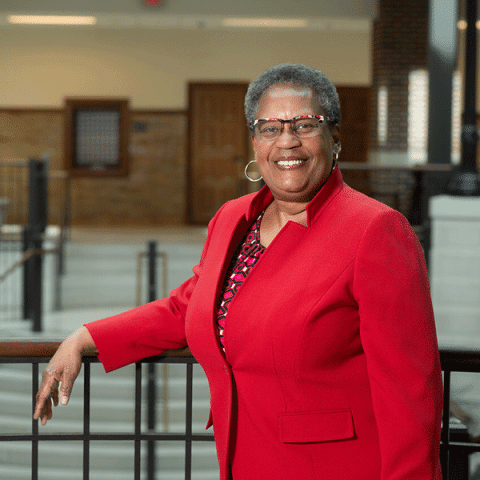 A Black woman with short gray hair and glasses stands smiling, wearing a bright red blazer over a patterned top. One hand rests on a railing, and a building interior with stairs and warm lighting is visible in the background.