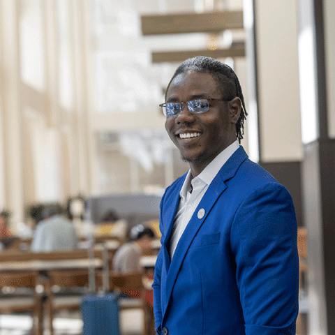 A young Black man with short locs and glasses, wearing a bright blue suit jacket and a white shirt, stands smiling indoors. People are seated and working in the background of the open, well-lit space.