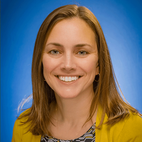 A white woman with straight light brown hair smiles in front of a blue background. She is wearing a yellow cardigan over a patterned top.