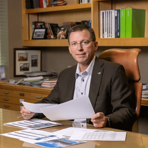 A white man with short brown hair and glasses sits at a desk holding documents. He is wearing a dark blazer over a patterned shirt. Papers are spread out in front of him, and shelves with books and framed photos are visible in the background.