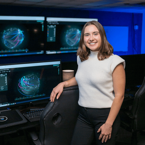 A white woman with shoulder-length light brown hair stands in a computer lab with multiple large monitors behind her displaying colorful digital data visualizations. She is smiling, wearing a light sleeveless sweater and dark pants, with one hand resting on a desk chair.