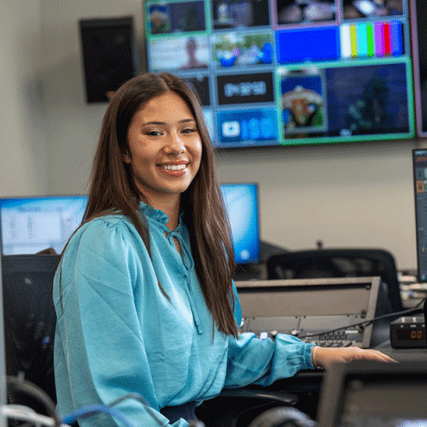 A young woman with long dark hair, wearing a light blue blouse, sits smiling at a control desk in a media production room, with multiple screens and monitors in the background.