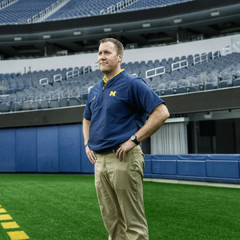 Four men stand together on a football field at night, smiling and celebrating. They are wearing matching white jackets and navy pants with an “M” logo, and two of them are holding roses. Confetti is falling in the background, and stadium lights and a large crowd are visible behind them.