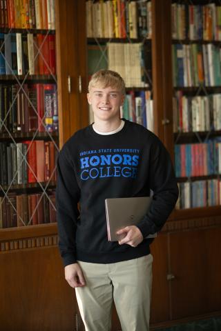A white male with short blond hair stands in front of a large wooden bookshelf filled with colorful books. He is wearing a black sweatshirt with the words “Indiana State University Honors College” printed in blue on the front, along with khaki dress pants. He is holding a closed laptop in one hand.