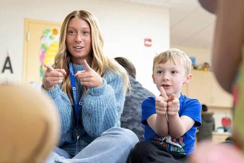 A white female with long blond hair wearing a blue sweater and jeans sits cross-legged on the floor next to a child in a blue T-shirt. Both are indoors in a classroom setting and are making hand gestures, pointing forward. The background shows a door with colorful decorations and other children seated nearby.