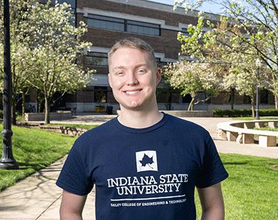 “A young man with short blond hair smiling while standing outdoors on a university campus, wearing a navy blue Indiana State University T-shirt. Trees, grass, and campus buildings are visible in the background.”