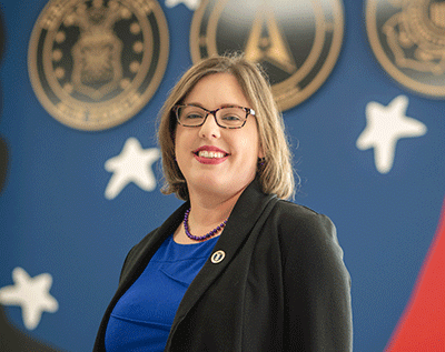 A person wearing glasses and a dark blazer over a blue top, smiling while standing indoors in front of a blue wall with decorative symbols and emblems.”
