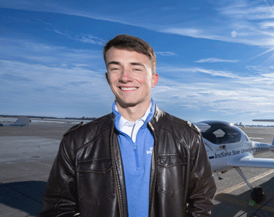 A smiling young white man with short brown hair, wearing a brown leather jacket over a blue shirt, standing on an airport tarmac with a small airplane and blue sky in the background.”