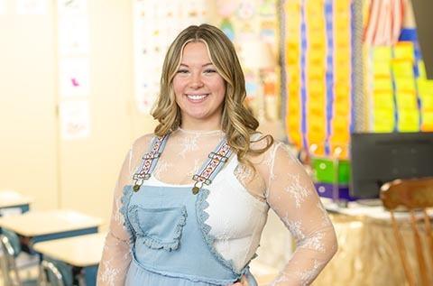 Person standing in a brightly decorated classroom, smiling at the camera while wearing a sheer long‑sleeve top and a light blue overall‑style dress, with desks, posters, and a bulletin board in the background.