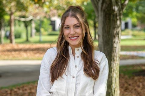“Portrait of a woman wearing a light-colored blouse, smiling outdoors with trees and a walkway in the background.”