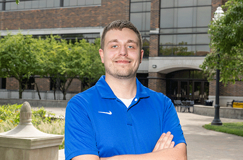“Portrait of a man in a blue polo shirt standing outdoors with his arms crossed, with trees and a walkway in the background.”