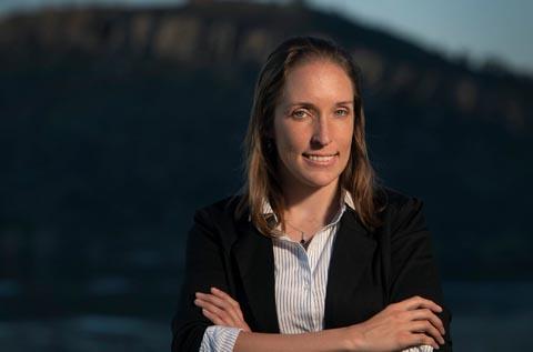 Professional portrait of a person standing outdoors with arms crossed, wearing a black blazer over a white and blue striped shirt, in front of a softly blurred mountainous landscape at sunset.