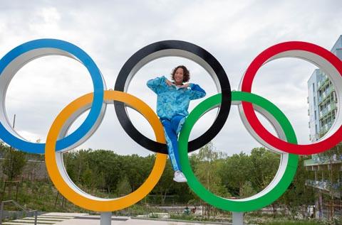 "Person sitting inside the black ring of a large outdoor Olympic rings sculpture, wearing a blue outfit and white shoes, posing with hands forming a heart shape. Trees and a building appear in the background."