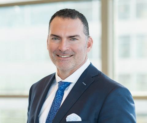 A white male with short brown hair wearing a navy suit with a blue patterned tie and white pocket square stands indoors in front of large windows in a modern office building.
