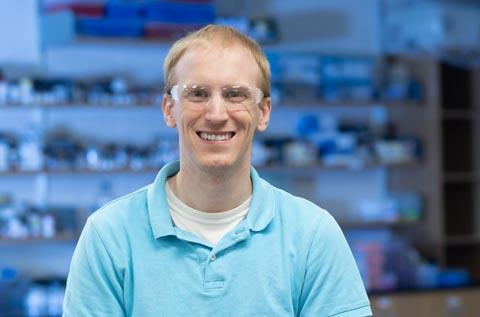 "Person wearing safety goggles and a light blue polo shirt seated in a laboratory, with shelves of supplies and equipment visible in the background."