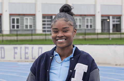 “Portrait of a woman wearing a letter jacket, smiling outdoors with a sports field in the background.”