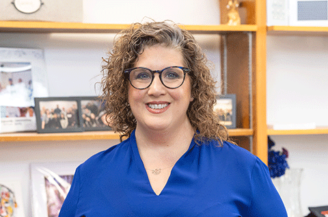 “Portrait of a woman wearing glasses and a blue top, smiling indoors with shelves in the background.”