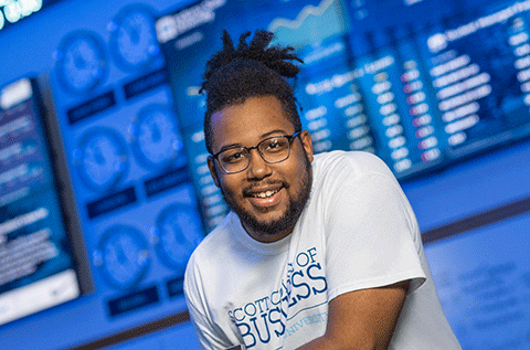“Portrait of a man wearing glasses and a white T-shirt, leaning forward and smiling, with digital screens visible in the background.”