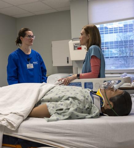 In a hospital room, two healthcare professionals stand beside a patient mannequin lying in a hospital bed, discussing care while medical equipment and a window with daylight are visible in the background.