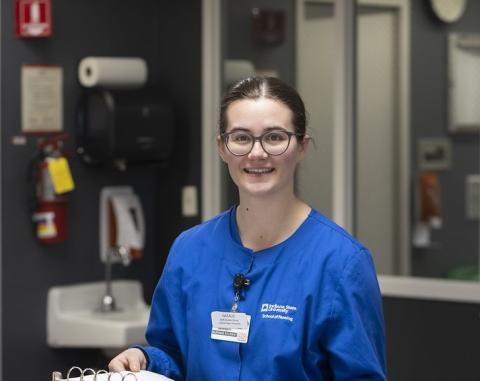 A white female student with brown hair pulled back wears blue nursing scrubs. She stands behind a reception desk in a healthcare setting, holding an open binder with documents, with sinks, cabinets, and medical supplies visible in the background.