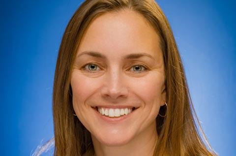 "Close-up portrait of a person smiling at the camera against a solid blue background, with long brown hair and small hoop earrings."