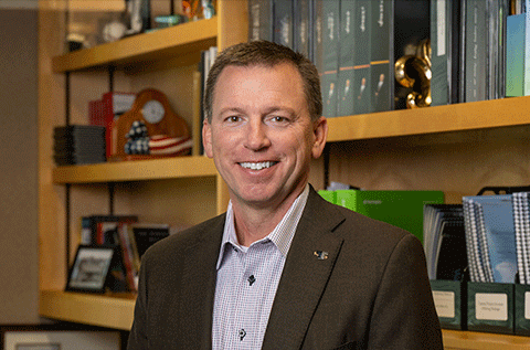 “Portrait of a man wearing a blazer and collared shirt, smiling indoors with bookshelves in the background.”