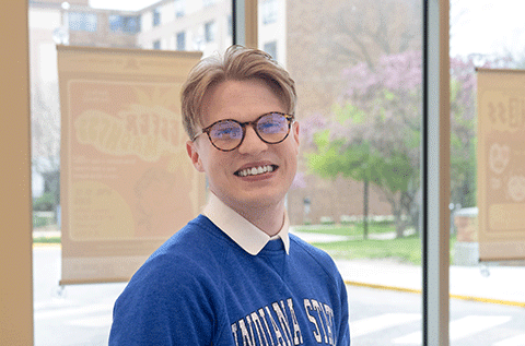 “Portrait of a man wearing glasses and a blue sweater, smiling indoors near a window.”