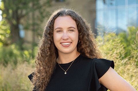 Portrait of a woman with curly hair wearing a black top, smiling outdoors with greenery in the background.”
