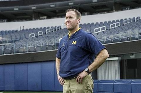 "Person standing in a stadium wearing a navy polo with a yellow “M” logo and khaki pants, hands on hips, with rows of empty seats in the background."