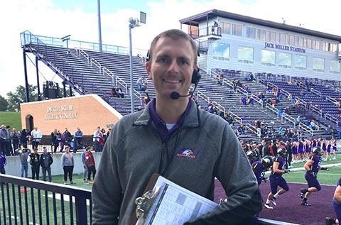 "Man wearing a headset and holding a clipboard while standing on a football field, with players practicing behind him and stadium seating visible in the background."