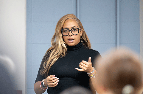 “Portrait of a woman wearing glasses and a black top, speaking and gesturing indoors, with people seated in the foreground.”