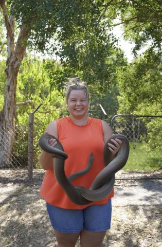 A white female student with blond hair pulled back wearing an orange sleeveless top and denim shorts holds a large dark-colored snake outdoors, with trees, fencing, and greenery in the background.