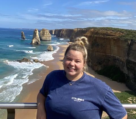 A white female with blond hair pulled back wearing a blue T-shirt stands on a cliff overlooking a sandy beach with turquoise waves, rocky formations, and coastal cliffs below.