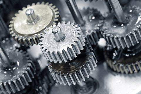 Close-up view of interlocking metal gears coated with lubricant, showing multiple toothed wheels mounted on shafts inside a mechanical assembly. The image emphasizes precision components and motion within machinery.