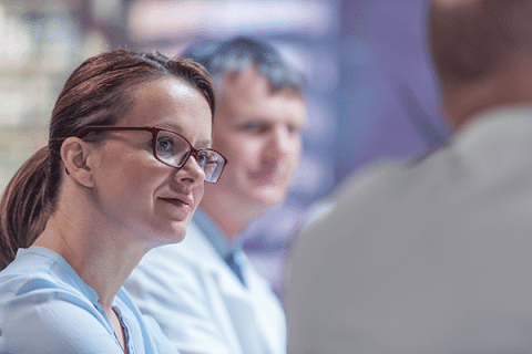 Healthcare professional wearing glasses listens attentively during a discussion with colleagues in a clinical setting.