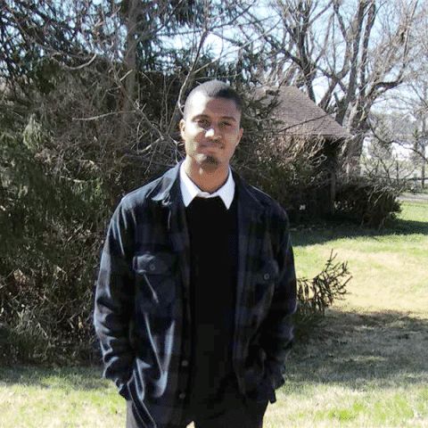 A Black male student standing outdoors on a grassy lawn, wearing a dark jacket over a sweater and collared shirt, with trees and a house visible in the background.