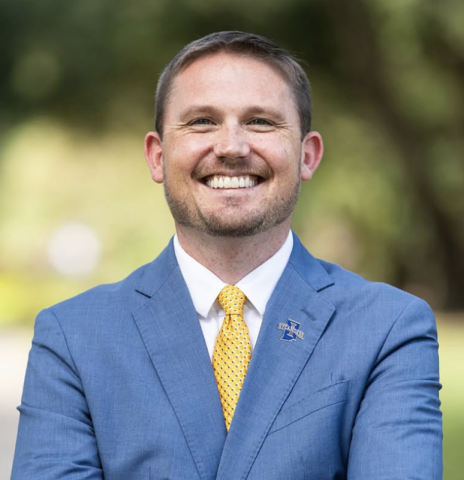 A white male with short brown hair stands outside wearing a blue suit, white undershirt, and a yellow tie. His arms are crossed over his chest. He smiles and poses for a headshot.