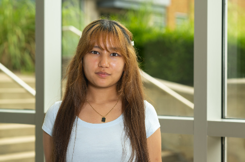 Female student with dark brown hair standing indoors near large windows in a campus building. The person is wearing a light blue shirt and a necklace, with the building exterior and greenery visible through the windows behind them.