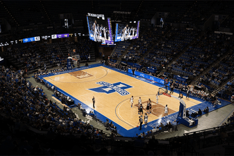 Wide view of an indoor basketball arena during a game, showing players on the court, spectators seated around the venue, and a large center-hung scoreboard above the floor. The court features a large “Sycamores” logo at center court, and the scoreboard displays a close score between Indiana State and UNI in the second half.