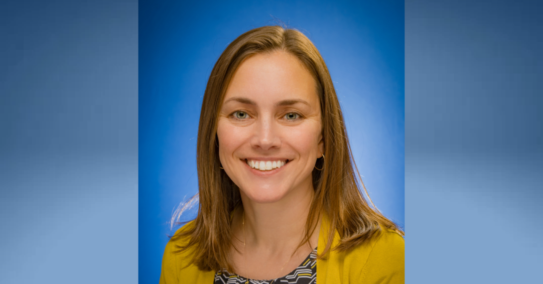 A white female with shoulder-length blonde hair, wearing a yellow cardigan over a patterned top, is posed in front of a blue background