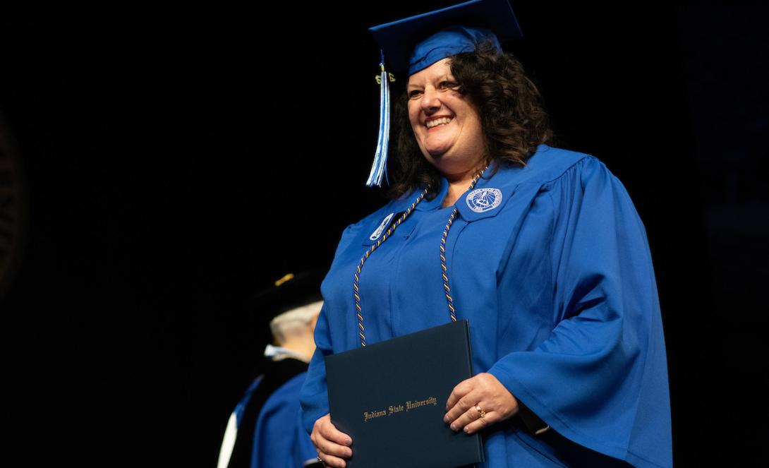 A white female with shoulder-length dark brown hair wearing a blue graduation gown and cap holds a diploma that reads “Indiana State University.”