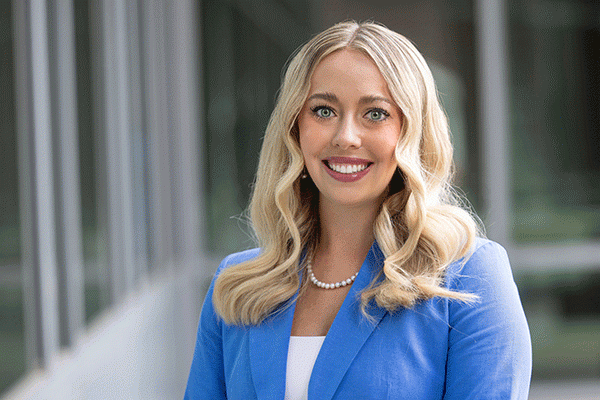 A smiling woman with long, wavy blonde hair wearing a blue blazer, a white top, and a pearl necklace. The background is an indoor setting with glass windows and blurred elements.