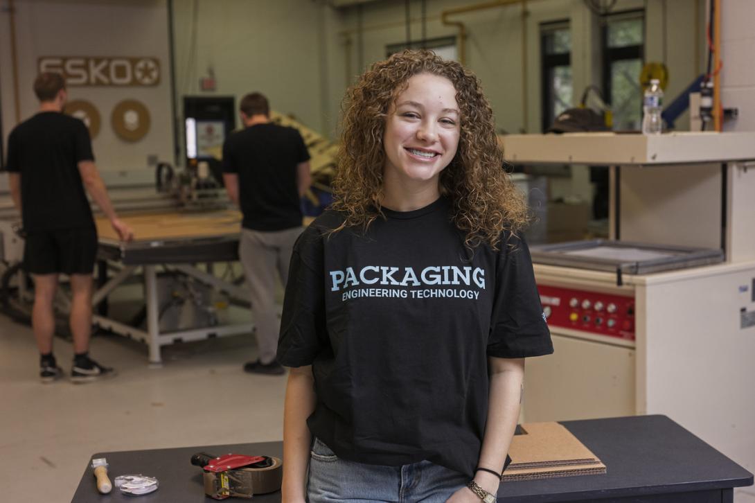 A light-skinned female student with curly brown hair wearing a black “Packaging Engineering Technology” T-shirt stands in a workshop with industrial equipment and worktables. Other students work in the background near machinery and tools.