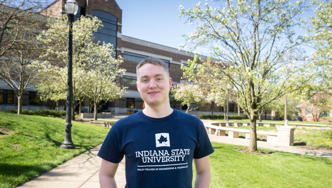 A light-skinned male student wearing an Indiana State University Bailey College of Engineering and Technology T-shirt stands outdoors on a sunny day in front of a brick academic building, surrounded by green grass, blossoming trees, and a walkway with benches.